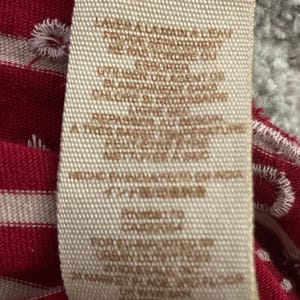 May include: A close-up of a clothing tag with care instructions in French. The tag is beige with brown text and is set against a red and white striped fabric. The fabric has white embroidered details.