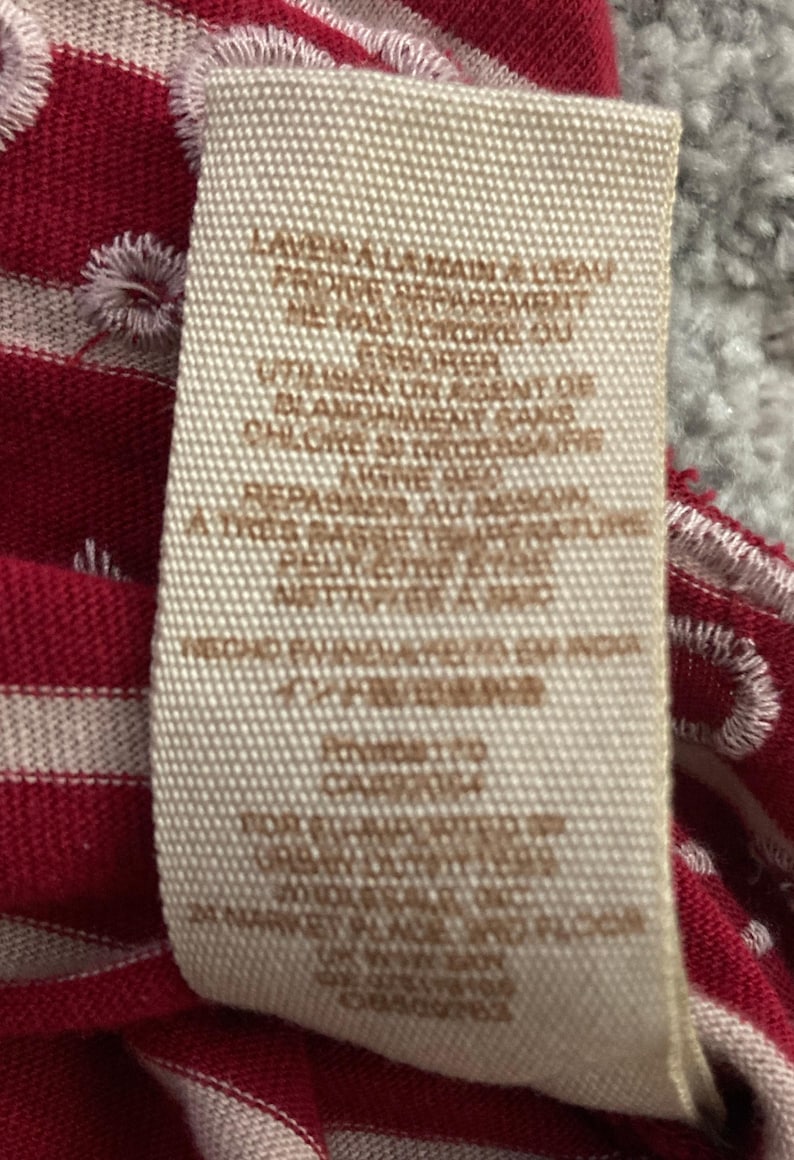 May include: A close-up of a clothing tag with care instructions in French. The tag is beige with brown text and is set against a red and white striped fabric. The fabric has white embroidered details.