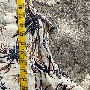 Puede incluir: Top blanco con estampado floral y ribete de encaje, medido con una cinta m&eacute;trica amarilla. La parte superior presenta un dise&ntilde;o bot&aacute;nico con acentos azul marino, naranja y marr&oacute;n. La parte superior mide aproximadamente 71 cm de largo.