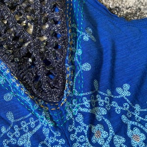 K&ouml;nnte beinhalten: Nahaufnahme eines blauen Oberteils mit H&auml;kel-Ausschnitt. Das Oberteil hat gestickte Blumenmuster in Hellblau und Wei&szlig;. Der Ausschnitt ist ein dunkelblauer H&auml;kelstoff mit silbernen Akzenten und dunkelblauen Perlen.