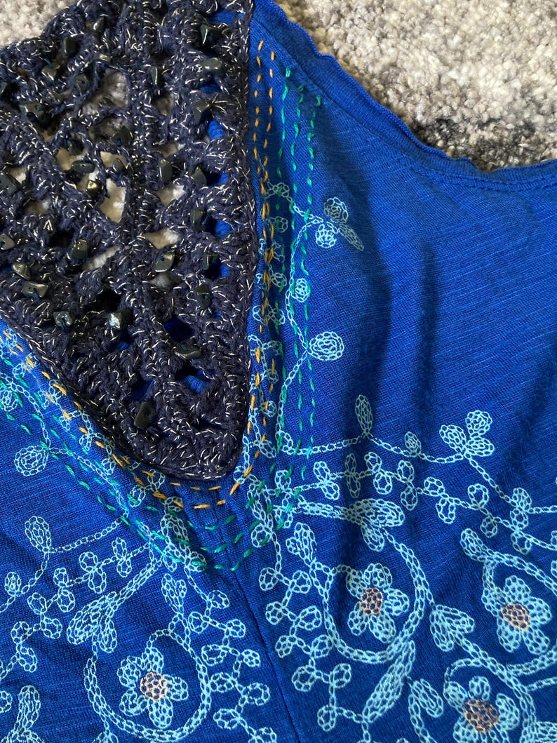 K&ouml;nnte beinhalten: Nahaufnahme eines blauen Oberteils mit H&auml;kel-Ausschnitt. Das Oberteil hat gestickte Blumenmuster in Hellblau und Wei&szlig;. Der Ausschnitt ist ein dunkelblauer H&auml;kelstoff mit silbernen Akzenten und dunkelblauen Perlen.