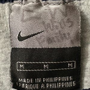 Pu&ograve; includere: Primo piano di un'etichetta di abbigliamento Nike. L'etichetta &egrave; grigia con un logo Nike swoosh nero. L'etichetta recita "MADE IN PHILIPPINES" e "FABRIQUE AUX PHILIPPINES". L'etichetta ha anche tre indicatori di taglia "M".