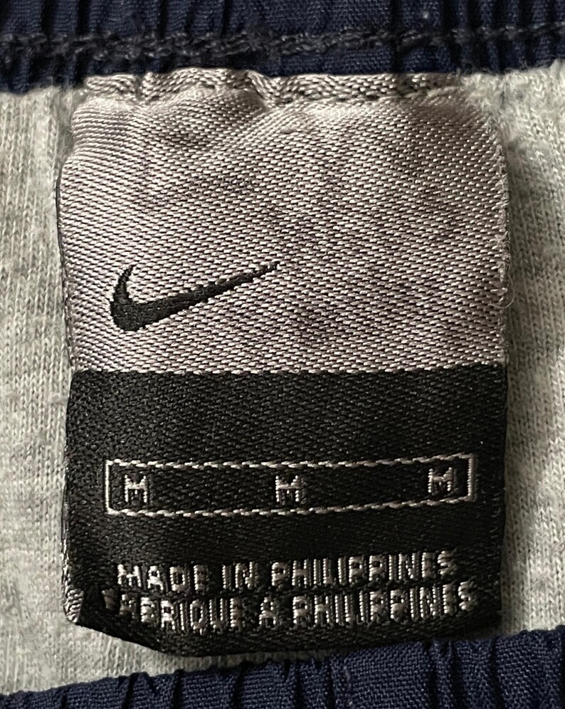 Pu&ograve; includere: Primo piano di un'etichetta di abbigliamento Nike. L'etichetta &egrave; grigia con un logo Nike swoosh nero. L'etichetta recita "MADE IN PHILIPPINES" e "FABRIQUE AUX PHILIPPINES". L'etichetta ha anche tre indicatori di taglia "M".