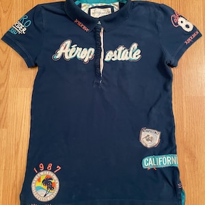 May include: A navy blue short-sleeve polo shirt featuring the embroidered text Aéropostale across the chest, a ruffled placket, and various colorful patches including a palm tree graphic, the number 8, and the word California.