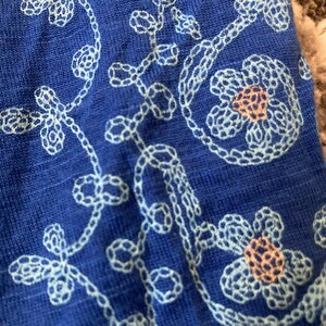 K&ouml;nnte beinhalten: Nahaufnahme eines blauen Stoffes mit wei&szlig;en, gestickten Blumen- und Rankenmustern. Die Stickerei zeigt detaillierte Blumen mit pfirsichfarbenen Zentren und zarten, geschwungenen Ranken, die ein strukturiertes Design bilden.