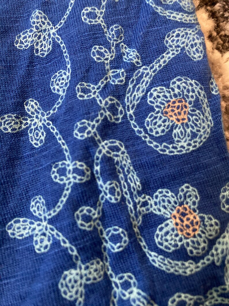 K&ouml;nnte beinhalten: Nahaufnahme eines blauen Stoffes mit wei&szlig;en, gestickten Blumen- und Rankenmustern. Die Stickerei zeigt detaillierte Blumen mit pfirsichfarbenen Zentren und zarten, geschwungenen Ranken, die ein strukturiertes Design bilden.