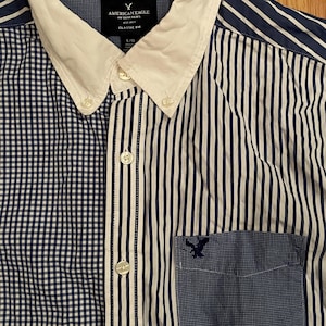 May include: A long-sleeve button-down shirt featuring a white collar and a mixed pattern design. The left side is blue and white gingham, while the right side and sleeves are blue and white striped. A blue gingham pocket features an eagle logo.
