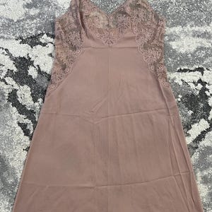May include: A dusty rose slip dress featuring delicate lace trim along the V-neckline, side panels, and hem. The garment includes thin, adjustable shoulder straps and a smooth, solid-colored fabric body that falls to a straight hemline.