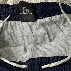 Pu&ograve; includere: Primo piano di pantaloncini sportivi blu navy e grigi con coulisse bianca. I pantaloncini hanno un pannello centrale grigio e finiture blu navy. &Egrave; visibile un'etichetta nera con il logo Nike e testo.