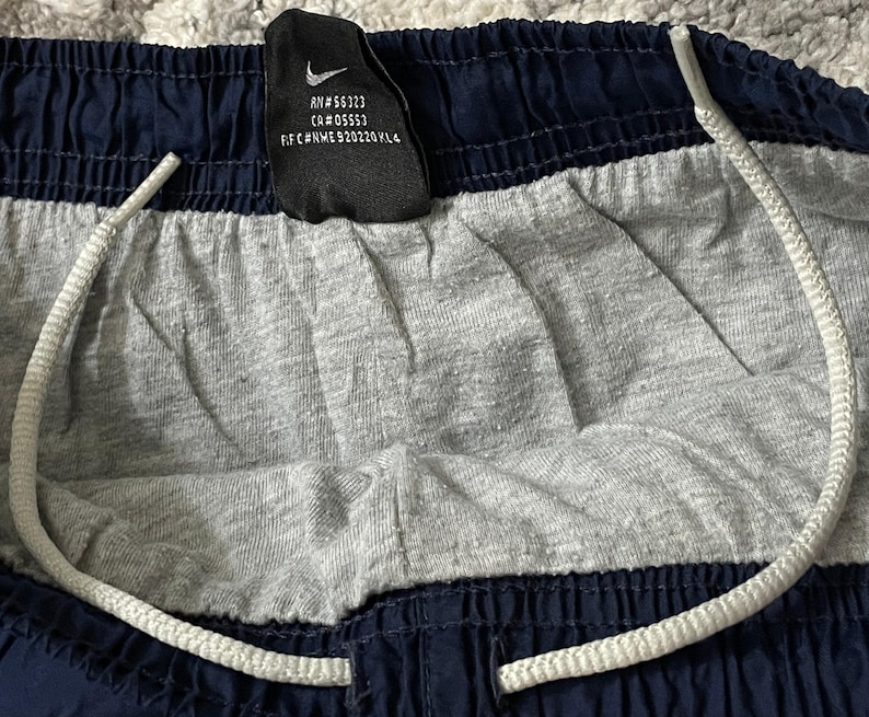 Pu&ograve; includere: Primo piano di pantaloncini sportivi blu navy e grigi con coulisse bianca. I pantaloncini hanno un pannello centrale grigio e finiture blu navy. &Egrave; visibile un'etichetta nera con il logo Nike e testo.