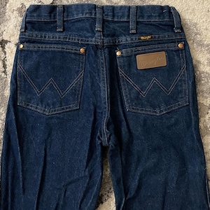 May include: Dark blue denim jeans with the Wrangler logo on the back pocket. The jeans have a classic five-pocket design with copper-colored rivets and a brown leather patch. The stitching is a contrasting tan color.