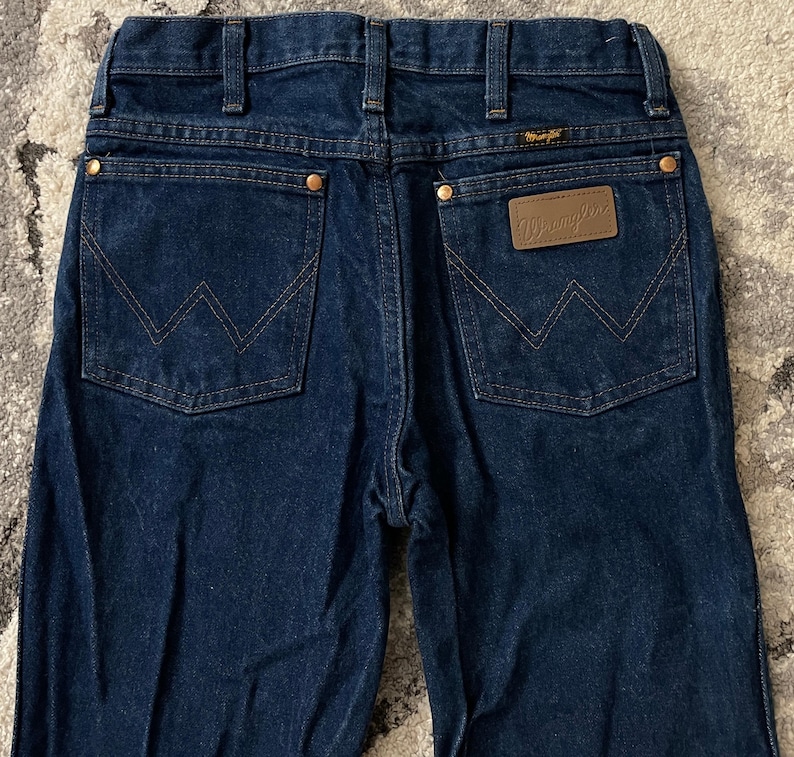 May include: Dark blue denim jeans with the Wrangler logo on the back pocket. The jeans have a classic five-pocket design with copper-colored rivets and a brown leather patch. The stitching is a contrasting tan color.