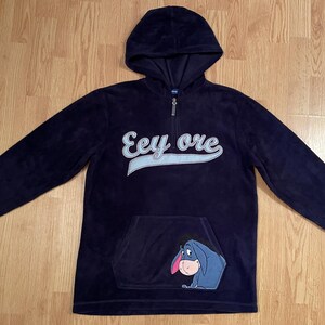 May include: A dark navy blue fleece hoodie featuring a quarter-zip neckline and long sleeves. The chest displays the text Eeyore in white script with a light blue underline. A graphic of the character Eeyore is positioned on the front pocket.