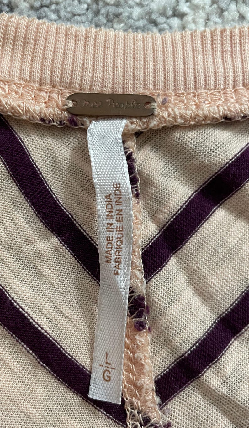 May include: Close-up of a light pink and cream-colored top with dark purple diagonal stripes. The top has a ribbed neckline and a tag that reads "Free People" and "Made in India / Fabriqu&eacute; en Inde".