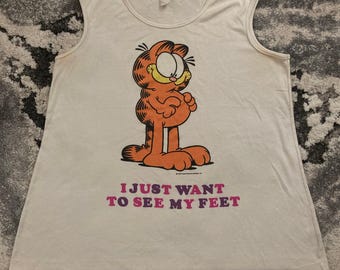 May include: Cream-colored sleeveless shirt featuring a cartoon Garfield character in orange. The graphic includes the text "I JUST WANT TO SEE MY FEET" in pink and purple. The shirt has a round neckline and a relaxed fit.