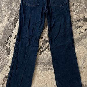 May include: Dark blue denim jeans with the Wrangler logo on the back pocket. The jeans have a classic five-pocket design and a straight leg fit. The jeans are laid flat on a patterned surface.