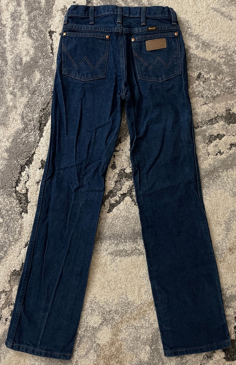 May include: Dark blue denim jeans with the Wrangler logo on the back pocket. The jeans have a classic five-pocket design and a straight leg fit. The jeans are laid flat on a patterned surface.