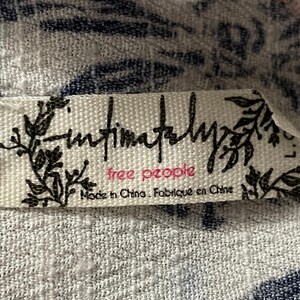 Puede incluir: Primer plano de una etiqueta de tela blanca con el texto "Intimately Free People" en escritura negra y letras rosas. La etiqueta tambi&eacute;n incluye el texto "Made in China. Fabriqu&eacute; en Chine" en negro. La etiqueta est&aacute; sobre una tela estampada blanca y azul.
