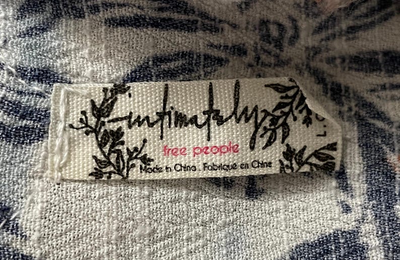 Puede incluir: Primer plano de una etiqueta de tela blanca con el texto "Intimately Free People" en escritura negra y letras rosas. La etiqueta tambi&eacute;n incluye el texto "Made in China. Fabriqu&eacute; en Chine" en negro. La etiqueta est&aacute; sobre una tela estampada blanca y azul.