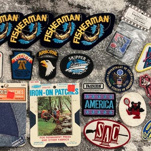 Könnte beinhalten: Eine Sammlung von aufgestickten Aufnähern mit verschiedenen Designs. Enthält Aufnäher mit den Wörtern "FISHERMAN", "AIRBORNE", "SKIPPER" und "AMERICA". Weitere Aufnäher zeigen Logos, Text und Illustrationen.