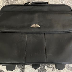 Puede incluir: Maletín rodante Samsonite negro con solapa superior y logotipo plateado. El maletín tiene un asa y dos ruedas en la parte inferior. El cuero muestra signos de desgaste.