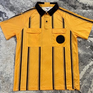 May include: A short-sleeved yellow polo shirt featuring vertical black stripes, a black collar, and a button-up placket. The shirt includes two chest pockets with flaps, and a solid black circular patch is attached to the left chest pocket.