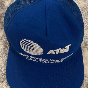 May include: A blue trucker hat with a mesh back and a curved brim. The hat features the AT&T logo in white, along with the text "Before Working Near Gas... CALL TOLL-FREE."