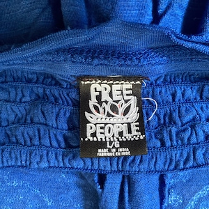 K&ouml;nnte beinhalten: Nahaufnahme eines leuchtend blauen Kleidungsst&uuml;cks mit ger&uuml;schtem Design. Ein schwarzes Etikett mit dem wei&szlig;en Text "FREE PEOPLE" und einem floralen Design ist angebracht. Das Etikett gibt auch die Gr&ouml;&szlig;e L/G und "MADE IN INDIA" an.