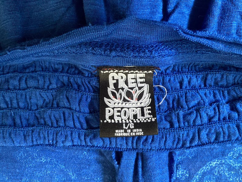 K&ouml;nnte beinhalten: Nahaufnahme eines leuchtend blauen Kleidungsst&uuml;cks mit ger&uuml;schtem Design. Ein schwarzes Etikett mit dem wei&szlig;en Text "FREE PEOPLE" und einem floralen Design ist angebracht. Das Etikett gibt auch die Gr&ouml;&szlig;e L/G und "MADE IN INDIA" an.