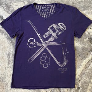 May include: A purple V-neck t-shirt with a white graphic of crossed tools, including scissors, a pipe wrench, and a crowbar. The shirt has a distressed design and the text "TABLE OF THE YEARS".