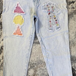 Op de afbeelding: Lichtblauwe spijkerbroek van verwassen denim met een hoge taille, knoop en ritssluiting. De linkerpijp heeft drie stoffen patches met gerafelde randen in de vorm van een roze driehoek, een gele cirkel en een oranje driehoek. De rechterpijp bevat een grote, rechthoekige patch met gerafelde randen die een gedetailleerde illustratie toont van een persoon in een spijkeroutfit met diverse kleurrijke grafische accenten.