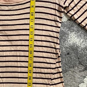 May include: A light pink long-sleeved shirt with horizontal dark brown stripes. The shirt is measured with a yellow measuring tape, showing a length of approximately 29 inches. The shirt has a scoop neckline.