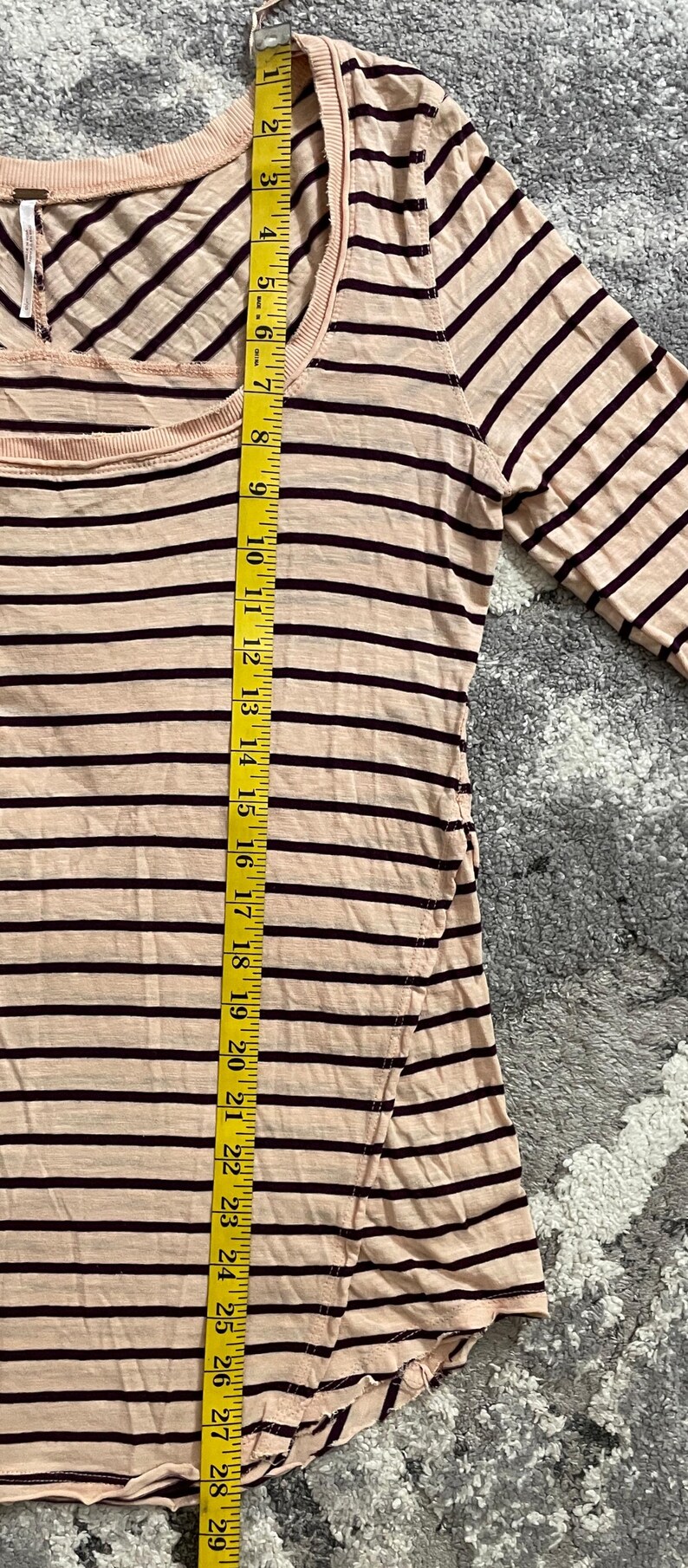 May include: A light pink long-sleeved shirt with horizontal dark brown stripes. The shirt is measured with a yellow measuring tape, showing a length of approximately 29 inches. The shirt has a scoop neckline.