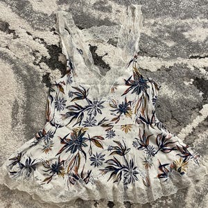 Puede incluir: Camisola blanca con estampado floral en azul marino, marr&oacute;n y amarillo. La parte superior presenta un ribete de encaje a lo largo del escote y el dobladillo, y una espalda de encaje. La camisola es un dise&ntilde;o sin mangas.