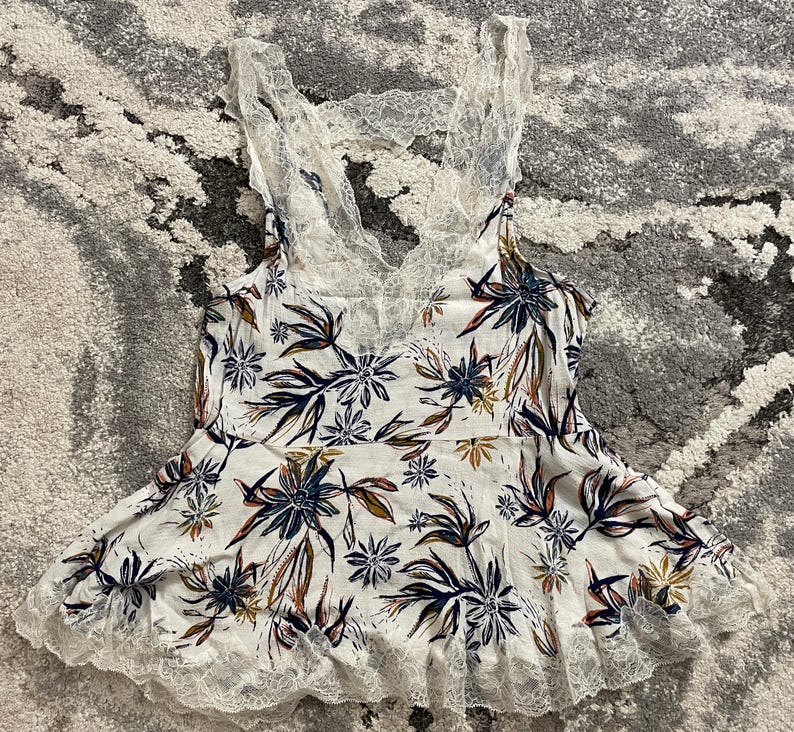 Puede incluir: Camisola blanca con estampado floral en azul marino, marr&oacute;n y amarillo. La parte superior presenta un ribete de encaje a lo largo del escote y el dobladillo, y una espalda de encaje. La camisola es un dise&ntilde;o sin mangas.