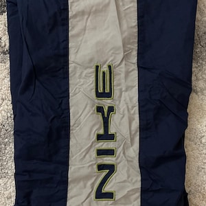 Pu&ograve; includere: Pantaloni sportivi blu navy e grigi con il marchio "NIKE" in verticale al centro in un font verde e giallo. I pantaloni sono realizzati in un materiale lucido, forse nylon.