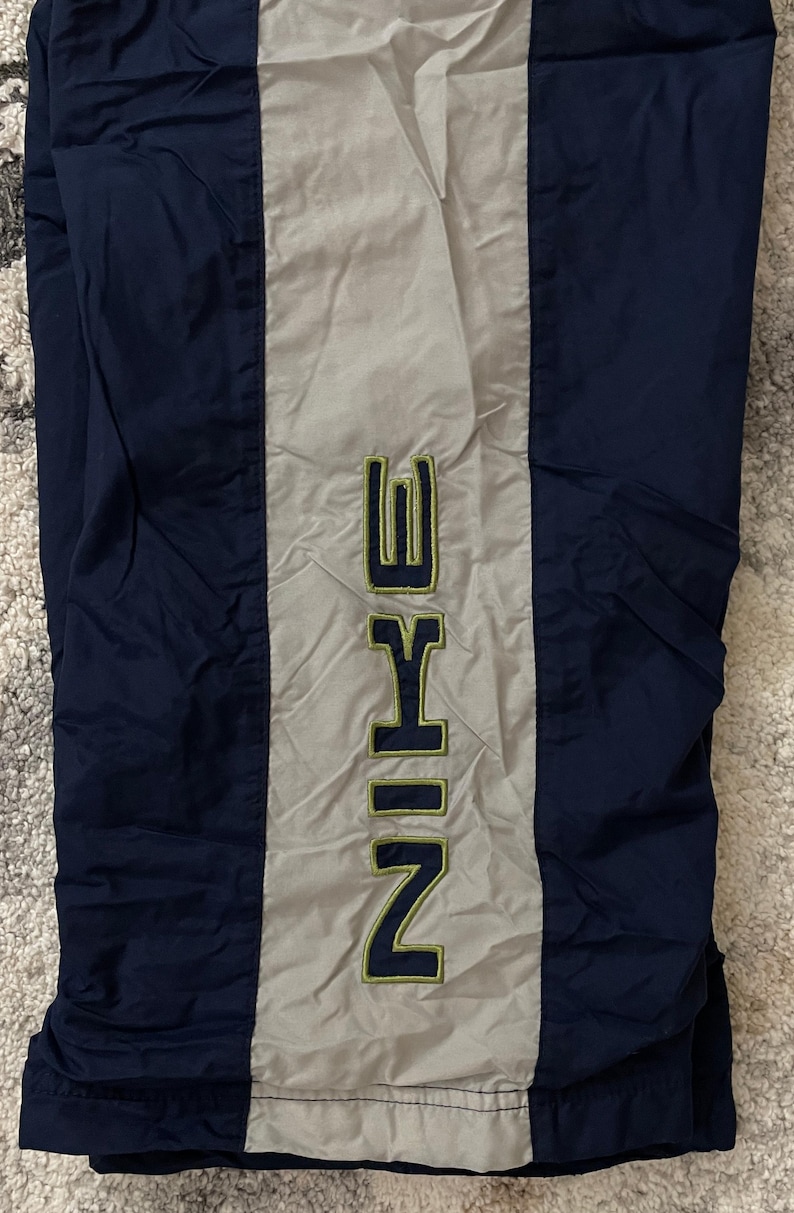 Pu&ograve; includere: Pantaloni sportivi blu navy e grigi con il marchio "NIKE" in verticale al centro in un font verde e giallo. I pantaloni sono realizzati in un materiale lucido, forse nylon.