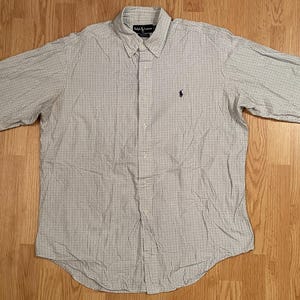 May include: A long-sleeve button-down shirt featuring a fine grid pattern in light blue and white. The shirt includes a pointed collar, a front button placket, and a small dark blue embroidered logo on the left chest area. The cuffs are buttoned.