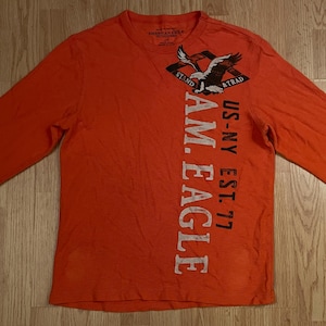 Peut inclure: Un t-shirt orange à manches longues avec un motif d'aigle au-dessus du texte "STAND & TRAD" et des lettres blanches verticales indiquant "AM. EAGLE" et "US-NY EST. 77". Le t-shirt présente un col rond et des poignets côtelés.