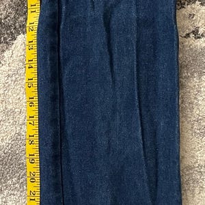 May include: Dark blue denim jeans with a straight leg, measured with a yellow tape measure. The jeans are approximately 32 inches long. The tape measure shows the length of the jeans.