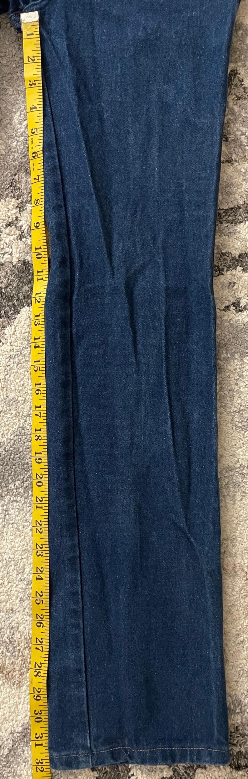 May include: Dark blue denim jeans with a straight leg, measured with a yellow tape measure. The jeans are approximately 32 inches long. The tape measure shows the length of the jeans.