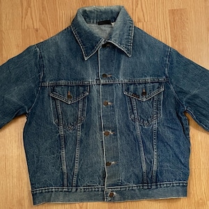 May include: A medium-wash blue denim jacket featuring a pointed collar, long sleeves with buttoned cuffs, and a front button closure. The garment includes two chest pockets with buttoned flaps and vertical seam detailing extending from the pockets.