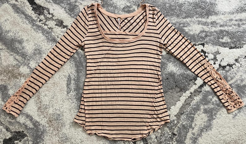 May include: A long-sleeved, scoop-neck shirt with horizontal black stripes on a light pink background. The sleeves have a lace detail at the wrist. The shirt is made of a soft, lightweight material.