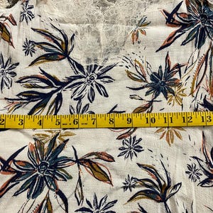 Puede incluir: Top blanco con estampado floral y escote de encaje. La tela presenta un estampado de flores y hojas azules, verdes y naranjas. Una cinta m&eacute;trica amarilla est&aacute; colocada sobre la tela.