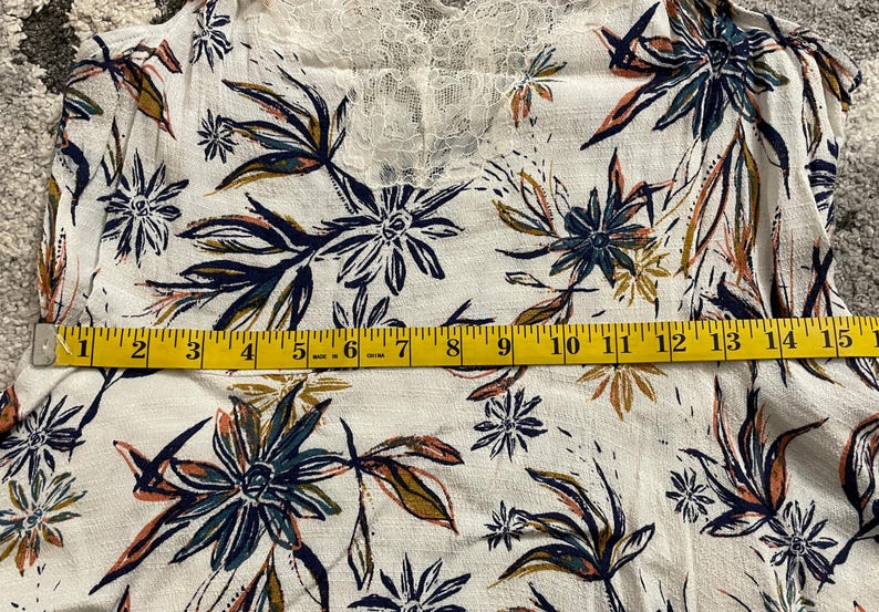 Puede incluir: Top blanco con estampado floral y escote de encaje. La tela presenta un estampado de flores y hojas azules, verdes y naranjas. Una cinta m&eacute;trica amarilla est&aacute; colocada sobre la tela.