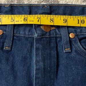 May include: Close-up of dark blue denim jeans with a yellow measuring tape across the waistband. The tape shows a measurement of approximately 14 inches. The jeans have copper-colored rivets and a button closure.