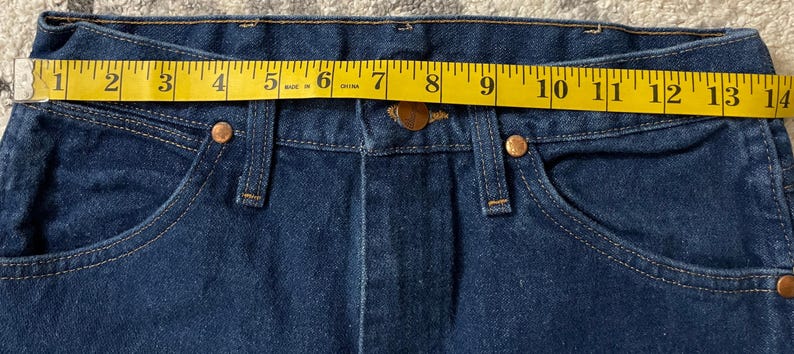 May include: Close-up of dark blue denim jeans with a yellow measuring tape across the waistband. The tape shows a measurement of approximately 14 inches. The jeans have copper-colored rivets and a button closure.