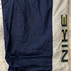 Pu&ograve; includere: Pantaloni sportivi blu navy e grigi con la scritta "NIKE" in lettere verdi. I pantaloni hanno una striscia verticale grigia sul lato e sono realizzati in un materiale leggero.