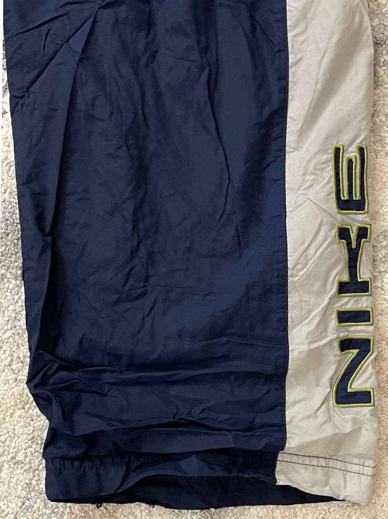Pu&ograve; includere: Pantaloni sportivi blu navy e grigi con la scritta "NIKE" in lettere verdi. I pantaloni hanno una striscia verticale grigia sul lato e sono realizzati in un materiale leggero.
