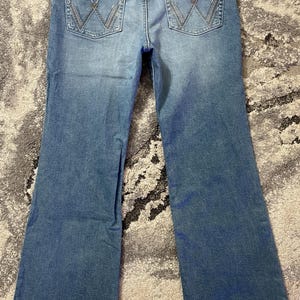 Puede incluir: Vaqueros de mezclilla azul claro con pernera acampanada. Los bolsillos traseros presentan un dise&ntilde;o de "W" cosido. Un parche de cuero marr&oacute;n con el logotipo "Wrangler" es visible en la cintura trasera.